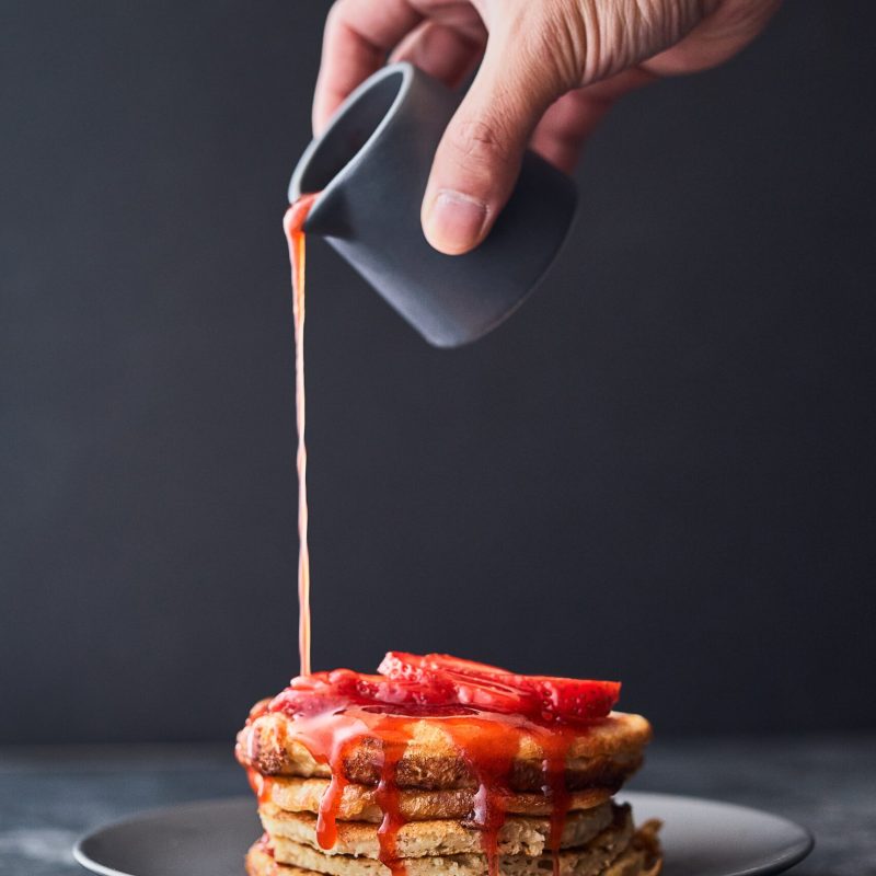 A vertical shot of a person pouring strawberry sauce on a stack of pancakes
